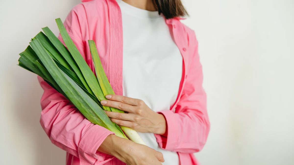 A woman in a pink jacket holding fresh spring onions against a white background.